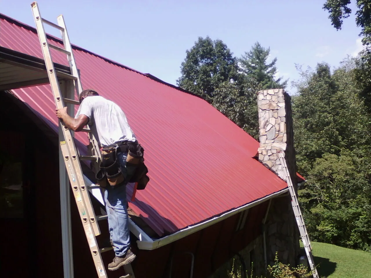Roofer on a ladder beside a red metal roof and stone chimney — Soffit & Fascia Repair by Roofing Brothers in Chestnut Mound